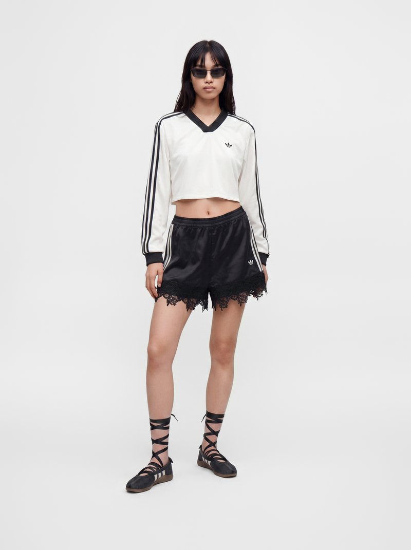 Γυναίκα που φοράει λευκό crop top μακρυμάνικο και μαύρα σορτς με δαντέλα, μαύρα παπούτσια με κορδόνια και σκούρα γυαλιά ηλίου, στέκεται μπροστά σε απλό φόντο.