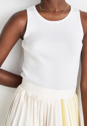 Vrouw die een witte mouwloze geribbelde top en een roomkleurige geplooide rok met lichtgele accentstrepen draagt, zichtbaar van nek tot taille.