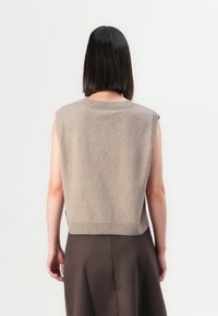 Persona con capelli neri lisci che indossa un top senza maniche beige a maglia e una gonna marrone scuro, in piedi di fronte a un muro bianco semplice.