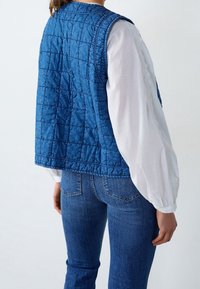 Blå vadderad väst med en lätt textur, med rund halsringning och ärmlös design, bärs över en långärmad vit blus.