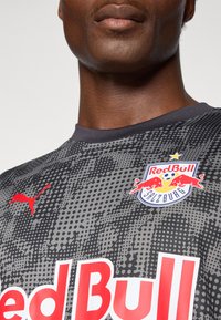 Maillot de sport gris avec un motif texturé, featuring le logo rouge de Puma et un grand texte "Red Bull". Emblèmes pour le Red Bull Salzbourg présents.