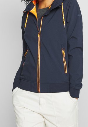 Person trägt eine marineblaue Jacke mit Reißverschluss, die einen leuchtend orangefarbenen Reißverschluss und Kordelzüge hat, kombiniert mit weißen Hosen, Hände in den Taschen.