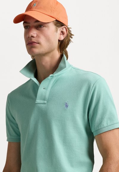 Lichtgroen polo shirt met een verhoogde kraag en een klein paars logo op de borst, gecombineerd met een oranje pet. Hulpige textuur.
