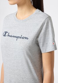 Szara bawełniana koszulka z krótkim rękawem z granatowym logo "Champion" na klatce piersiowej. Krótkie rękawy z małym akcentem logo na lewym rękawie.