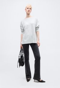 Γυναίκα που φοράει ανοιχτό γκρι πουλόβερ με ανοιχτούς ώμους, μαύρο τζιν, μαύρα ψηλοτάκουνα, κρατώντας μαύρη τσάντα σε λευκό, απλό φόντο.