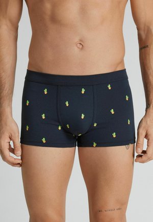 Męski tors w ciemnych bokserkach z wzorem małych zielonych kaktusów w pomarańczowych doniczkach, stojący z rękami swobodnie opuszczonymi wzdłuż ciała.