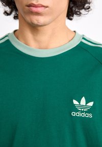 Žalias medvilninis marškinėliai su šviesiai žaliai raibota apykakle, turintis mažą baltą Adidas logotipą ir tris baltas juosteles ant pečių.
