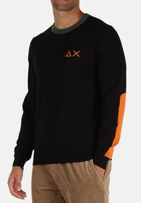Maglione nero con colletto a girocollo verde, presenta un logo arancione sul petto e un dettaglio arancione sulla manica sinistra. Texture morbida.