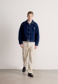 Mannens mørkeblå denimjakke med frontlommer, kontrastsøm, brukt over en lys grå skjorte, kombinert med beige bukser og svarte sko.