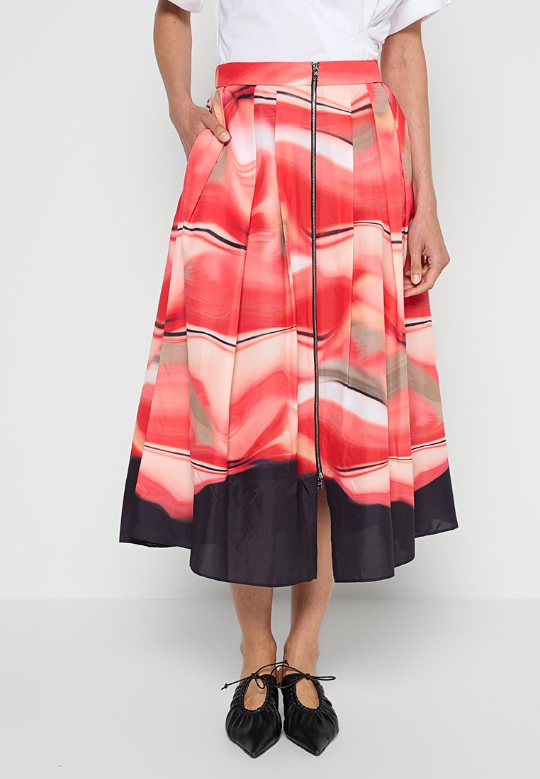 Kobieta nosząca plisowaną midi spódnicę w kolorze czerwonym, różowym i czarnym z zamkiem z przodu, białą bluzkę oraz czarne baleriny z zadartym noskiem i wiązaniami.