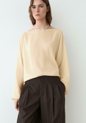 Frau trägt eine beige Langarmbluse, in hoch taillierte, plissierte dunkle Hose gesteckt, ergänzt durch eine durchsichtige Perlenkette.