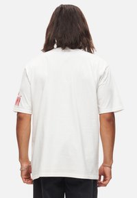 T-shirt blanc en coton avec des manches courtes, col rond et un graphique rouge sur la manche gauche. Texture lisse, coupe décontractée.