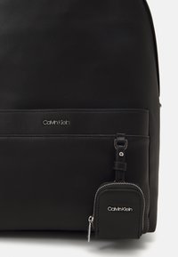 Black backpack made of textured nylon with a smooth leather accent. Features a front pocket and a detachable mini pouch with a zipper.