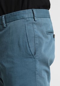 Pantaloni azzurri chiari realizzati in morbido tessuto, dotati di zip frontale, tasche laterali e una tasca posteriore con chiusura a bottone. Design slim fit.