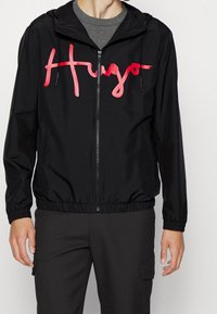 Black windbreaker jacket featuring a red "Hugo" logo, elastic cuffs and hem, and a hood with drawstrings. Smooth material, zippered front.