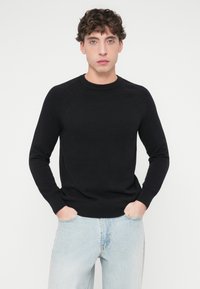 Μαύρο πλεκτό πουλόβερ με στρογγυλή λαιμόκοψη, ριμπ λεπτομέρειες στους ώμους και μακριά μανίκια. Συνδυασμένο με ανοιχτόχρωμο μπλε τζιν.