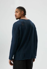 Donkerblauwe trui met lange mouwen en een textuurrijke afwerking, voorzien van meerdere verweerde stukken en een brede halslijn. Zichtbare naden en een ontspannen pasvorm.