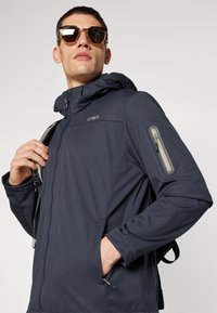 Navy blue soft shell jacket with a zippered chest pocket and hand pockets, adjustable hood, and logo detail on the front.