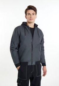 Jachetă bomber gri închis cu glugă, închidere cu fermoar, manșete elastice și buzunare laterale; țesătură texturată cu un luciu subtil și design sportiv.