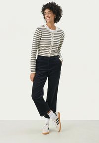 Gestreepte katoenen cardigan met knoopsluiting, gecombineerd met marineblauwe corduroy broek en witte sneakers met zwarte strepen. Model staat met de handen in de zakken.
