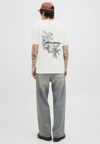 Crèmekleurig t-shirt met een kleurrafisch tropisch ontwerp op de achterkant, gecombineerd met lichtgrijze rechte jeans en zwarte schoenen.