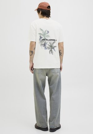 Crèmekleurig t-shirt met een kleurrafisch tropisch ontwerp op de achterkant, gecombineerd met lichtgrijze rechte jeans en zwarte schoenen.