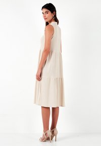Beige sleeveless dress with textured fabric, tiered design, knee-length, and subtle waistband. Paired with light beige heeled sandals.