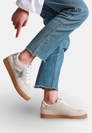 Personne portant des baskets beige clair avec des détails en mesh et un accent en forme d'étoile, associées à un jean bleu raccourci, pointant du doigt vers une chaussure.