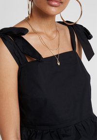 Svart klänning med fyrkantig halsringning, slipsar som knyts med en rosett och en samlad midja. Accessoarer inkluderar lager av guldhalsband och stora hoopörhängen.