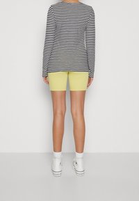 Osoba stojąca tyłem, ubrana w czarno-biały pasiasty top z długimi rękawami, żółte szorty, białe skarpety i białe trampki za kostkę.