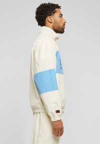 Lightweight, cream windbreaker with blue horizontal stripes, high collar, elastic cuffs, and embroidered logo detail on chest.