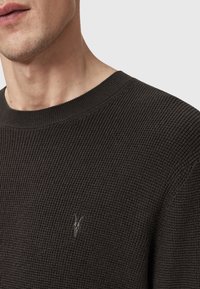 Lähikuva henkilöstä, joka käyttää tummanharmaata teksturoitua neulepaitaa, jossa on pieni brodeerattu logo vasemmassa rinnassa.
