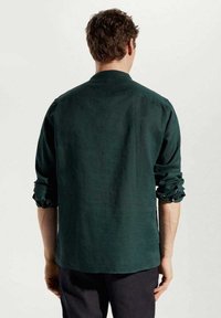 Camicia a maniche lunghe verde scuro realizzata in un tessuto testurizzato, con maniche arrotolate e orlo dritto, vista da dietro.