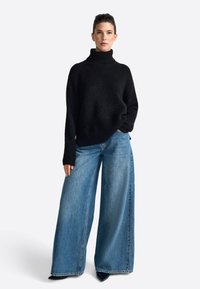 Fekete kötött garbós pulóver, laza szabással, világoskék, bő szárú farmernadrággal párosítva. A farmer nadrág klasszikus denim felületű és enyhén kopott.