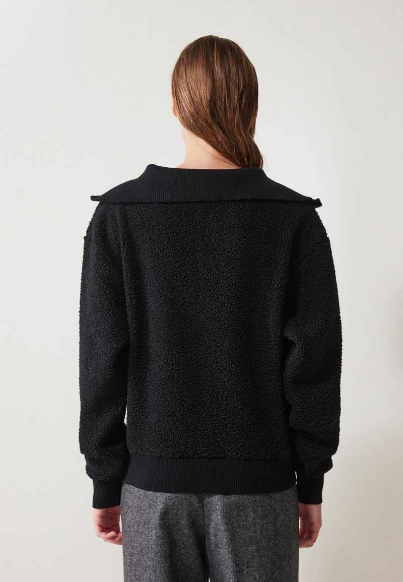 Pull noir texturé avec une finition en laine, présentant un large col et des poignets et une taille côtelés. La vue arrière montre une coupe décontractée.