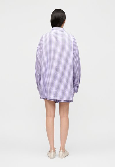 Γυναίκα με μακριά σκούρα μαλλιά στέκεται με την πλάτη, φορώντας υπερμεγέθη λιλά πουκάμισο με καρό και αντίστοιχα σορτς, με λευκά αθλητικά παπούτσια.