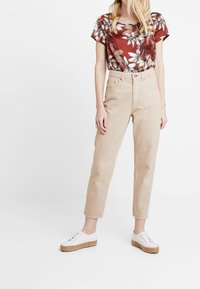 Rød blomstret kortærmet top med hvide og grønne mønstre, parret med højtaljede beige bukser og hvide sko med jute detaljer.