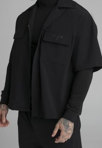 Svart kortärmad jacka med öppen front, två bröstfickor och ett mönstrat tyg. Bärs över en svart långärmad skjorta.