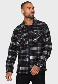 Camisa de franela a cuadros en negro, blanco y naranja, con dos bolsillos en el pecho, cierre de botones y una textura de madera, llevada sobre una parte superior negra.