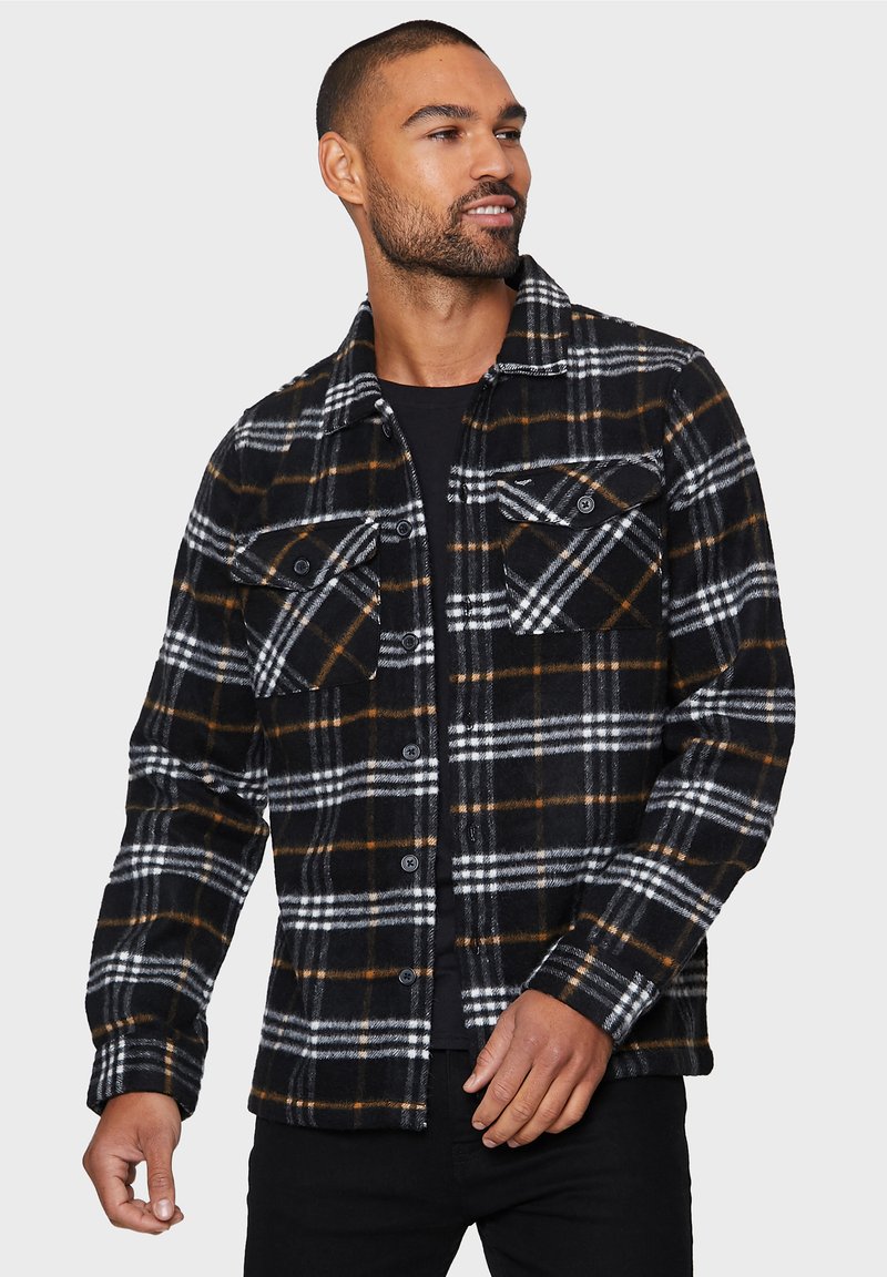 Camisa de franela a cuadros en negro, blanco y naranja, con dos bolsillos en el pecho, cierre de botones y una textura de madera, llevada sobre una parte superior negra.