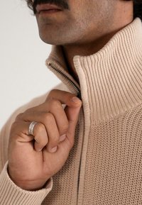 Nahaufnahme einer Person, die einen beigen Strickpullover mit hohem Kragen zippt, während sie einen silbernen strukturierten Ring an ihrer Hand trägt.