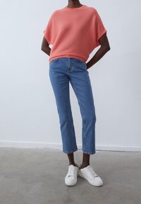 Korálový pletený oversized svetr spárovaný s modrými rovnými džíny a bílými teniskami, s uvolněným střihem a ležérním stylem.