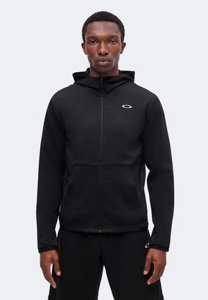 Man met zwarte zip-up hoodie en bijpassende zwarte broek met klein wit logo op borst en been, staand voor een effen achtergrond.