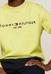 Žlutá mikina s kulatým výstřihem, černým nápisem "TOMMY HILFIGER EST. 1985", jemná textura a žebrované manžety, doplněná tmavými kalhotami.