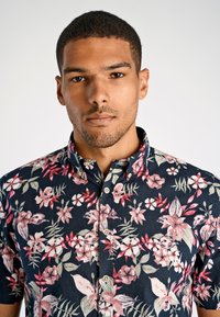 Granatowa koszula krótki rękaw w tropikalny wzór kwiatowy w odcieniach różu i zieleni, z kołnierzykiem zapinanym na guziki i fakturowanym materiałem.