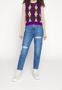 Chaleco de punto con un patrón argyle en púrpura, marrón y beige, combinado con jeans azules de tiro alto y rotos, y zapatillas beige con cordones.
