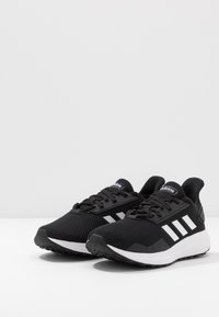 Czarne buty sportowe z siateczkową cholewką, trzema białymi paskami po boku i białą podeszwą środkową. Gumowa podeszwa zapewniająca przyczepność i trwałość.