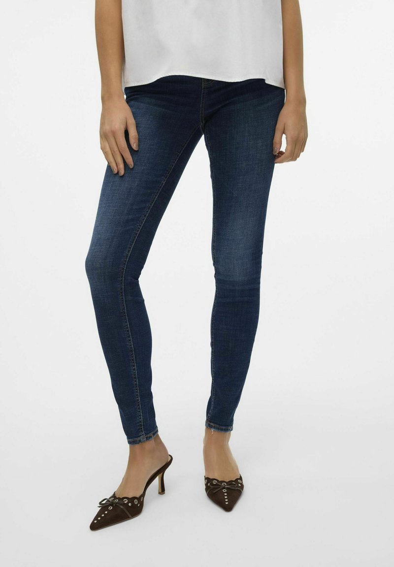 Slimfit jeans van donkerblauwe denim met een gladde textuur, gecombineerd met bruine puntige muiltjes met decoratieve accenten.