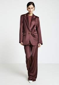 Frau trägt ein dunkel mauvefarbenes Satinsakko mit einem einzigen Knopf, passende weite Hosen und weiße spitze Pumps, steht auf einem weißen Boden.