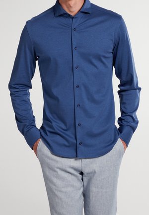 Uomo che indossa una camicia blu a maniche lunghe con colletto e pantaloni grigio chiaro, mani nelle tasche, su sfondo neutro.